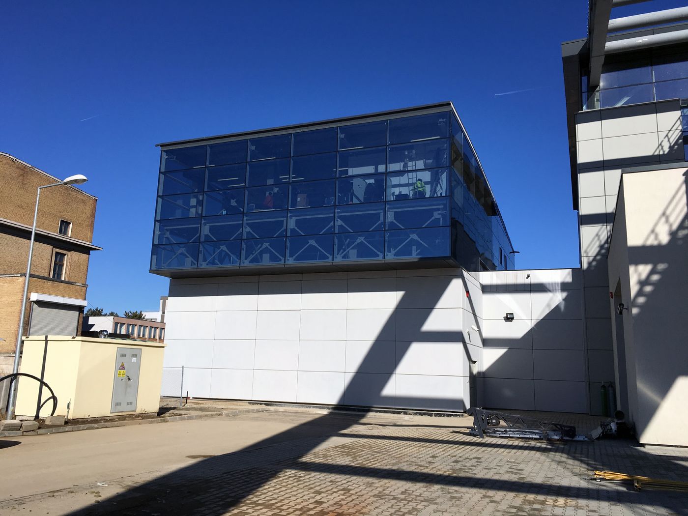 Photo: street-view of a building with steel structure, concrete and glass elements; on the left, electric appliances; on the right, part of another building