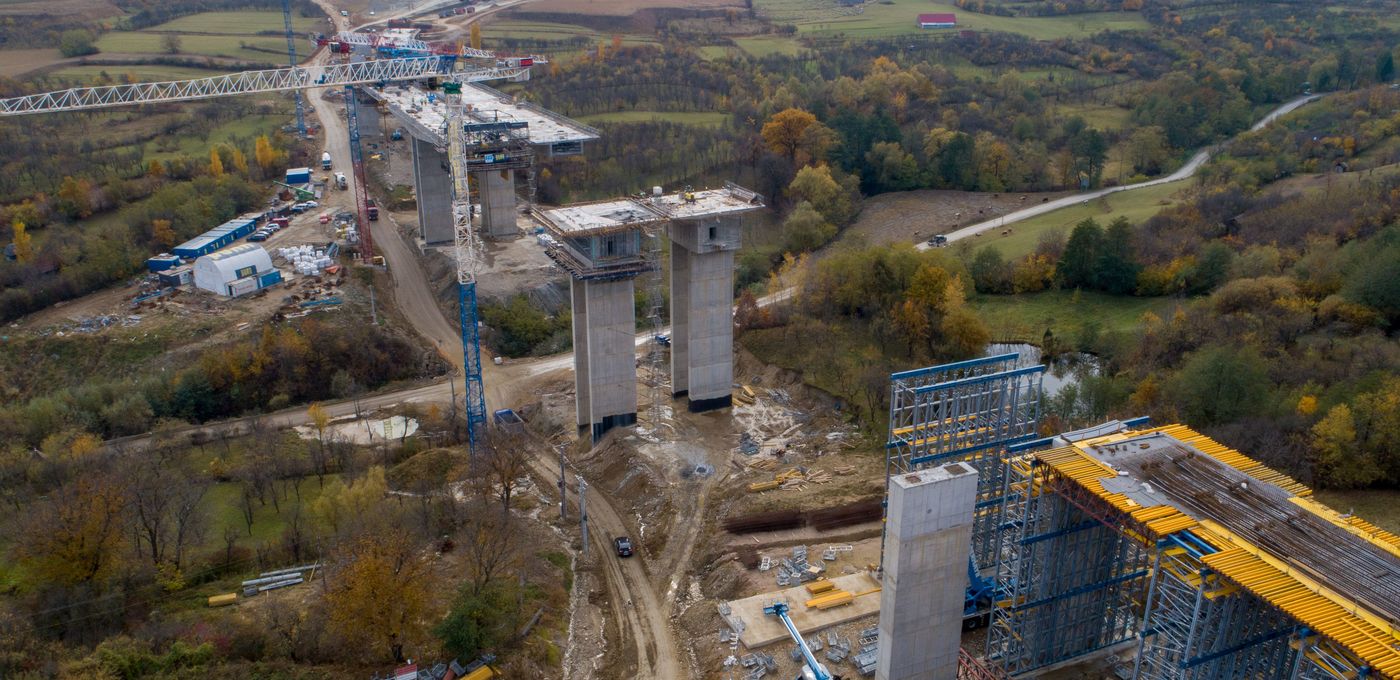 Aerial photo: pairs of still unconnected bridge piers, some of them scaffolded, and a construction crane amidst a rural area, mountains in the background.