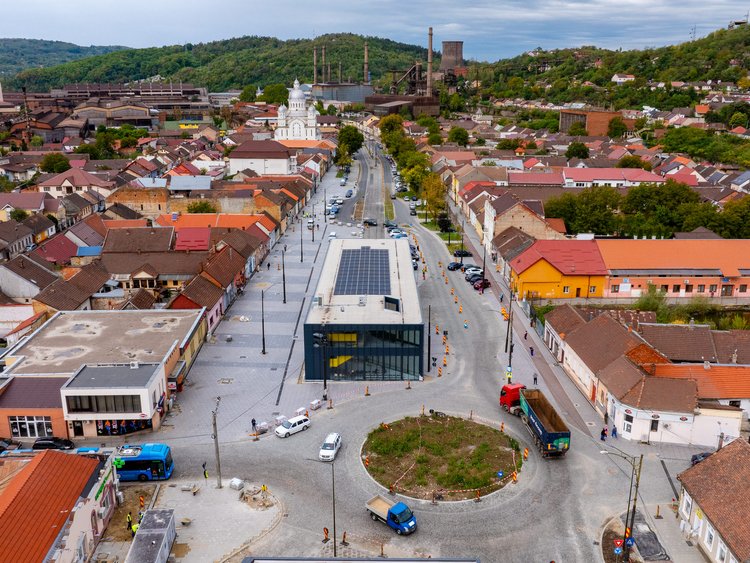For a greener future A slight bird's eye view of a modern tram terminus with photovoltaic modules on the roof. Newly laid wide pavements and roads around the station. In the foreground a roundabout, several vehicles are on the road. The station is surrounded by low houses, with wooded hills in the background.