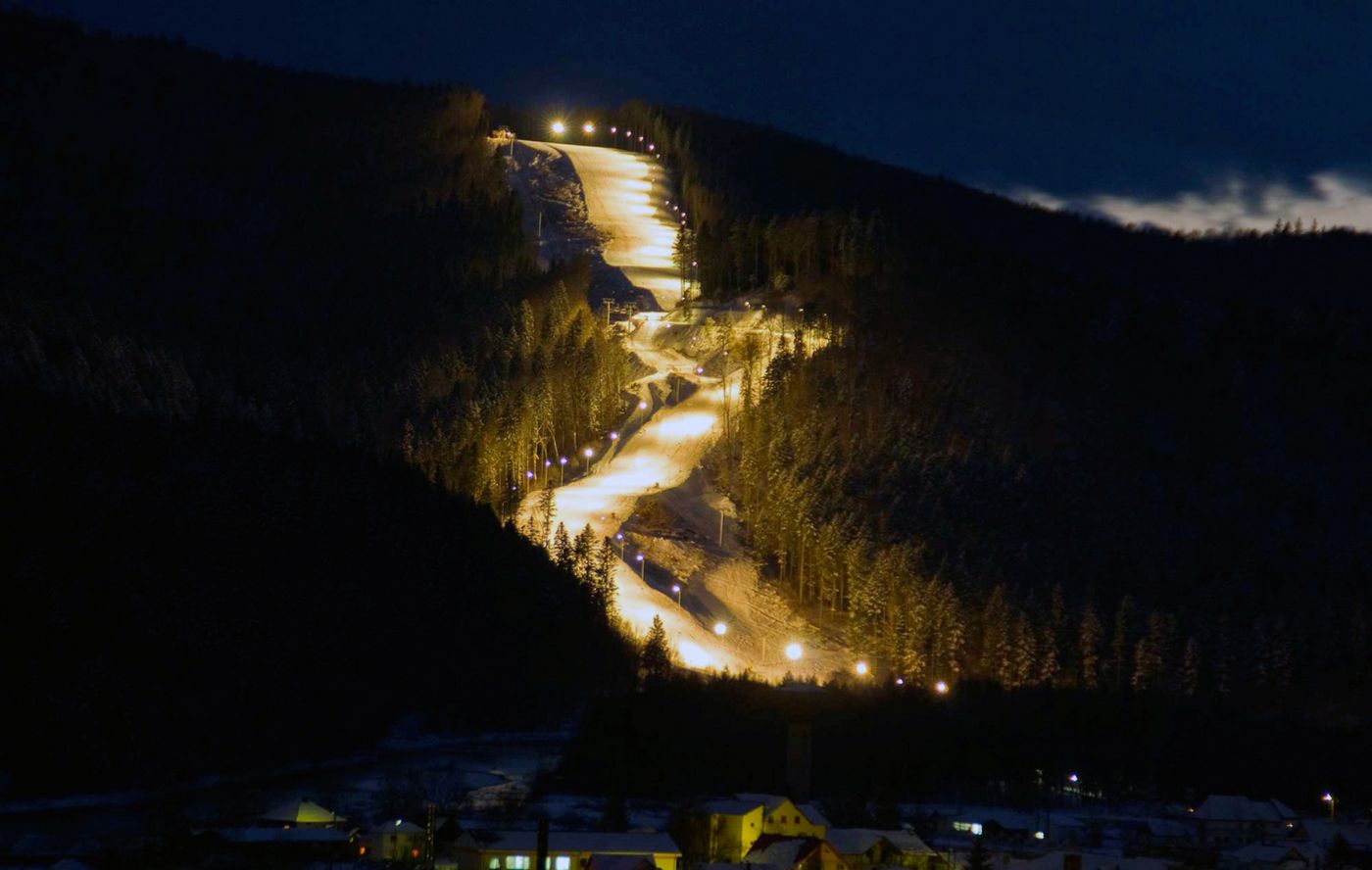 Photo: view of an illuminated ski slope by night; surrounded by forest; in the background, a dark blue night sky