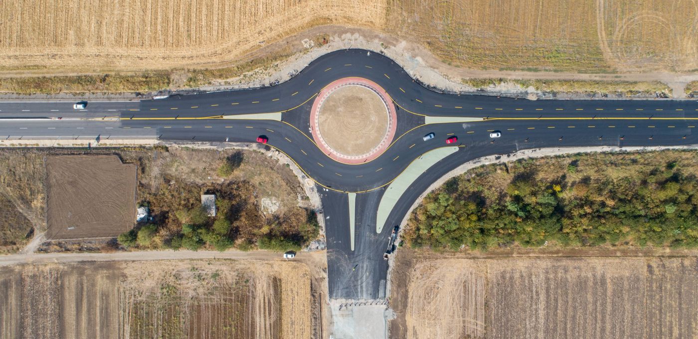 Aerial photo: direct view on a roundabout junction of roads to the left, right and down in amidst agricultural land, some cars driving on the road.