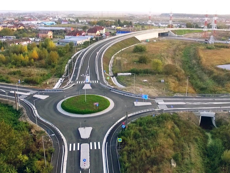 An example of professional construction work.  Photo: aerial view of four roads ending in a roundabout; the overpass in the background; surroundings of green grass, trees, some houses and parked cars