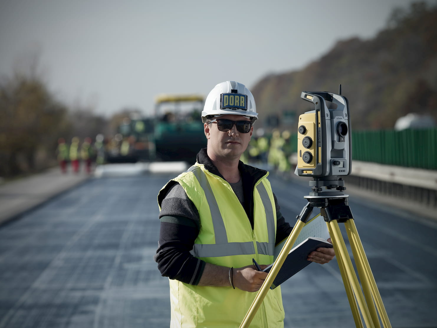 Photo: worker wearing safety vest, PORR helmet and sunglasses, holding a book for taking notes, next to an optical leveling instrument; blurred construction site with freshly cemented road in the background
